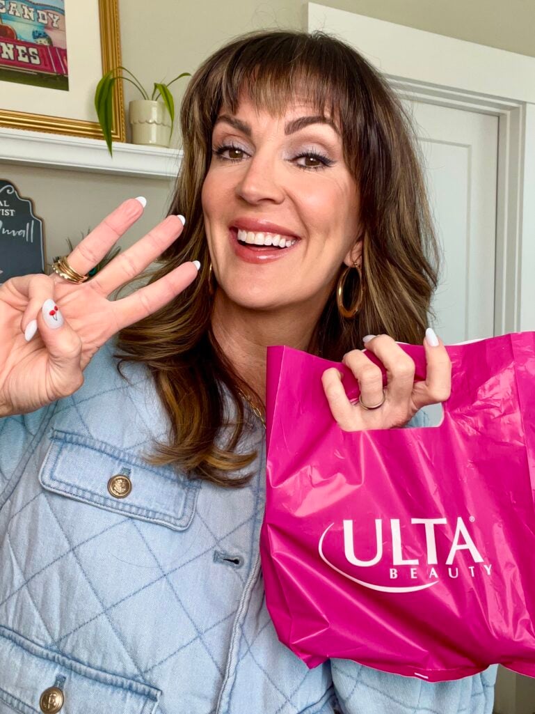 Woman with highlighted brown hair and bangs wearing quilted light-wash denim jacket, smiling and holding pink Ulta Beauty shopping bag while flashing a peace sign, cherry nail art on white nails, gold hoop earrings