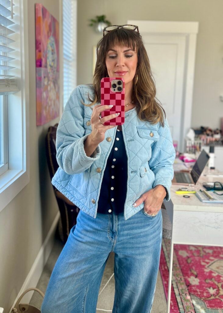 Full-length mirror selfie of woman in all-denim outfit — quilted light-wash denim jacket over navy square neck sweater and wide-leg light-wash jeans — holding red checkered phone case, tortoiseshell sunglasses on head, makeup vanity visible in background