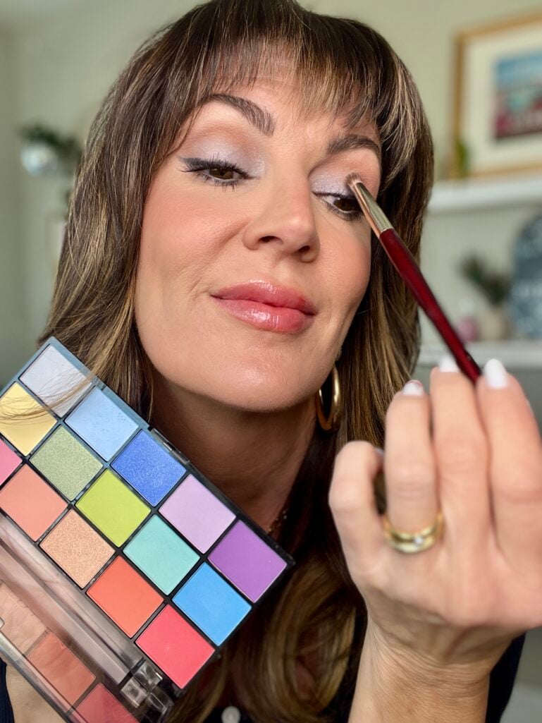 Close-up of woman applying blue denim eyeshadow with red-handled brush while holding colorful Revolution Reloaded Sugar Pie eyeshadow palette, gold hoop earrings, glossy neutral lip, white French manicure