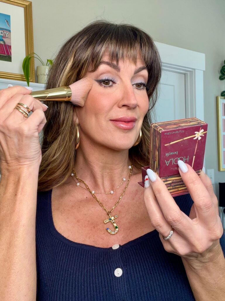 Woman applying Benefit Hoola bronzer to her cheek with a large gold-handled powder brush, holding the iconic Hoola compact, wearing navy square neck sweater and layered gold necklaces including a bejeweled J initial pendant