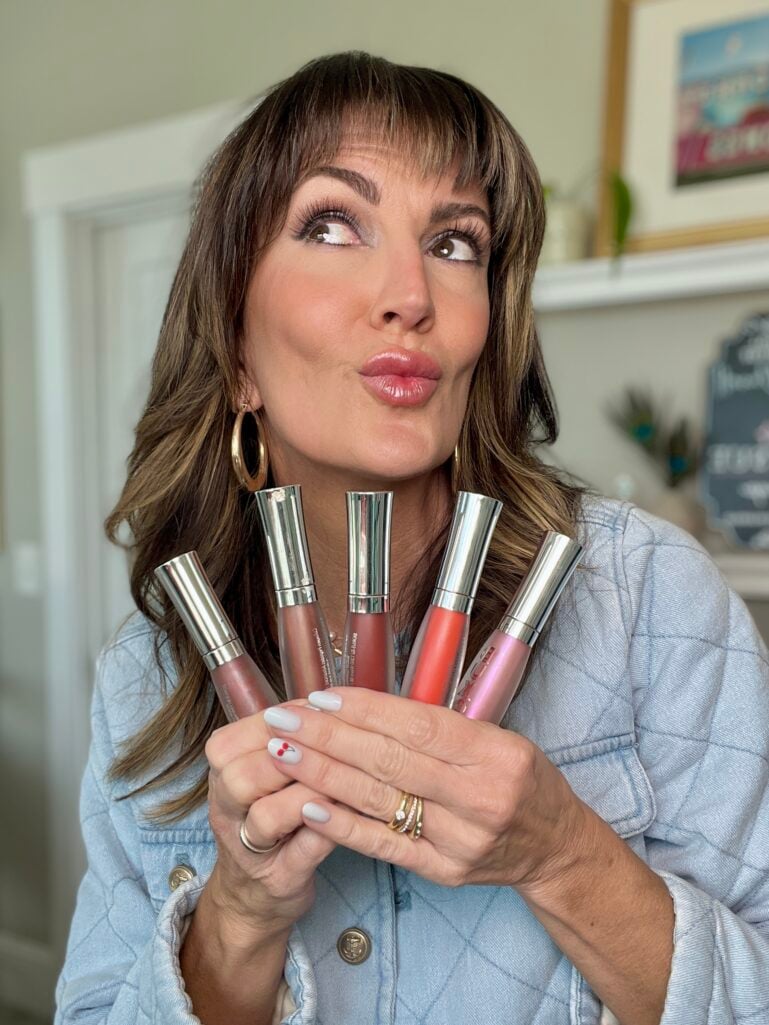 Woman in quilted denim jacket holding a fan of five Buxom Plump Shot Plumping Lip Serum Gloss tubes in assorted shades, making a pouty kiss face, gold hoop earrings, cherry nail art on white nails