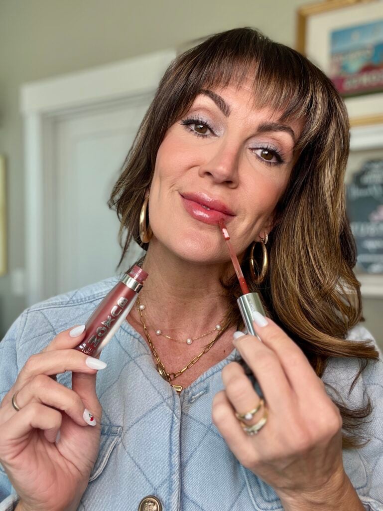 Woman applying Buxom lip gloss with the wand while holding two Buxom Plump Shot tubes, wearing quilted denim jacket and gold hoop earrings, blue denim eyeshadow and pearl chain necklace visible