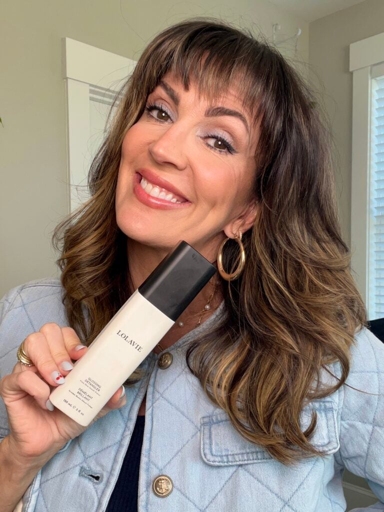 Woman smiling and holding a bottle of LolaVie Glossing Detangler hair product, wearing quilted light-wash denim jacket, gold hoop earrings, blue eyeshadow, highlighted brunette waves