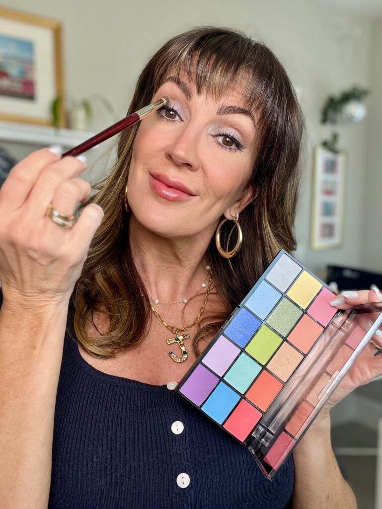 Woman applying blue eyeshadow with a red-handled brush while holding the Revolution Reloaded Sugar Pie colorful eyeshadow palette, wearing navy top and layered gold necklaces including a bejeweled J initial pendant, gold oval hoop earrings