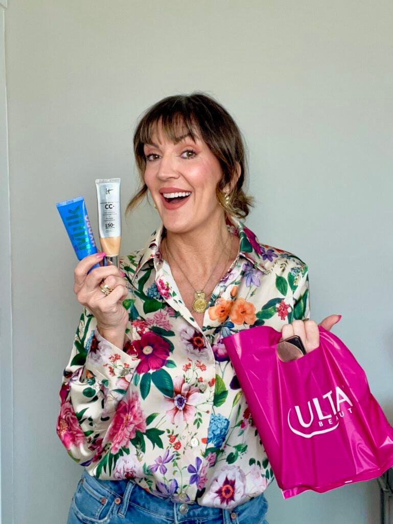 Woman with brown hair and bangs wearing silky ivory button-down blouse with bright floral print in pinks, purples, greens, and oranges, holding blue Milk Makeup Hydro Grip tube and It Cosmetics CC Cream tube in one hand and hot pink Ulta Beauty bag in the other, denim jeans, gold coin pendant necklace, excited expression, hot pink nails