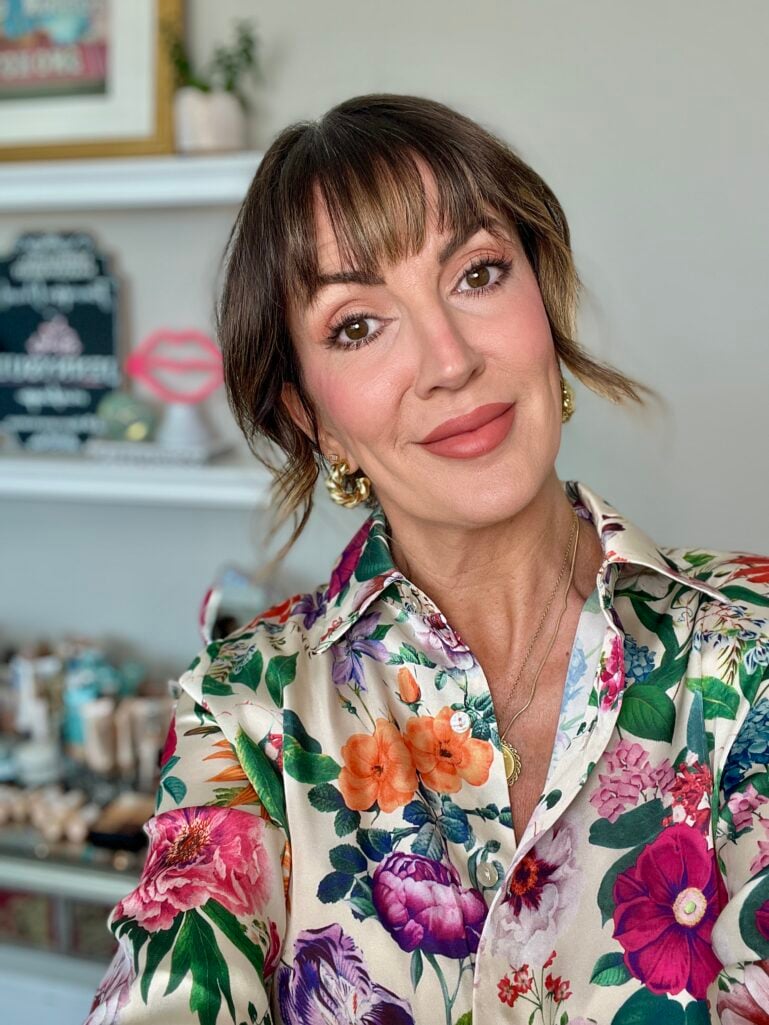 Close-up portrait of woman with brown hair pinned up and curtain bangs wearing Georgia Peach makeup look with warm peach eyeshadow, peachy-pink flushed cheeks, and matte peach lips, gold chunky hoop earrings, gold coin pendant necklace, silky floral blouse, beauty products on shelf in soft-focus background