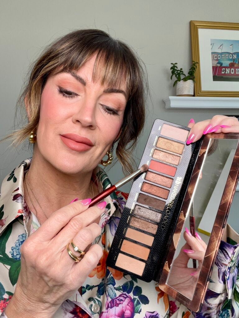 Woman with eyes partially closed holding Urban Decay Naked Reloaded eyeshadow palette open showing warm neutral and peach shimmer shades with built-in mirror, dipping eyeshadow brush into peach shade, floral blouse, hot pink nails, gold hoop earrings and rings, vintage cotton candy art print on shelf in background