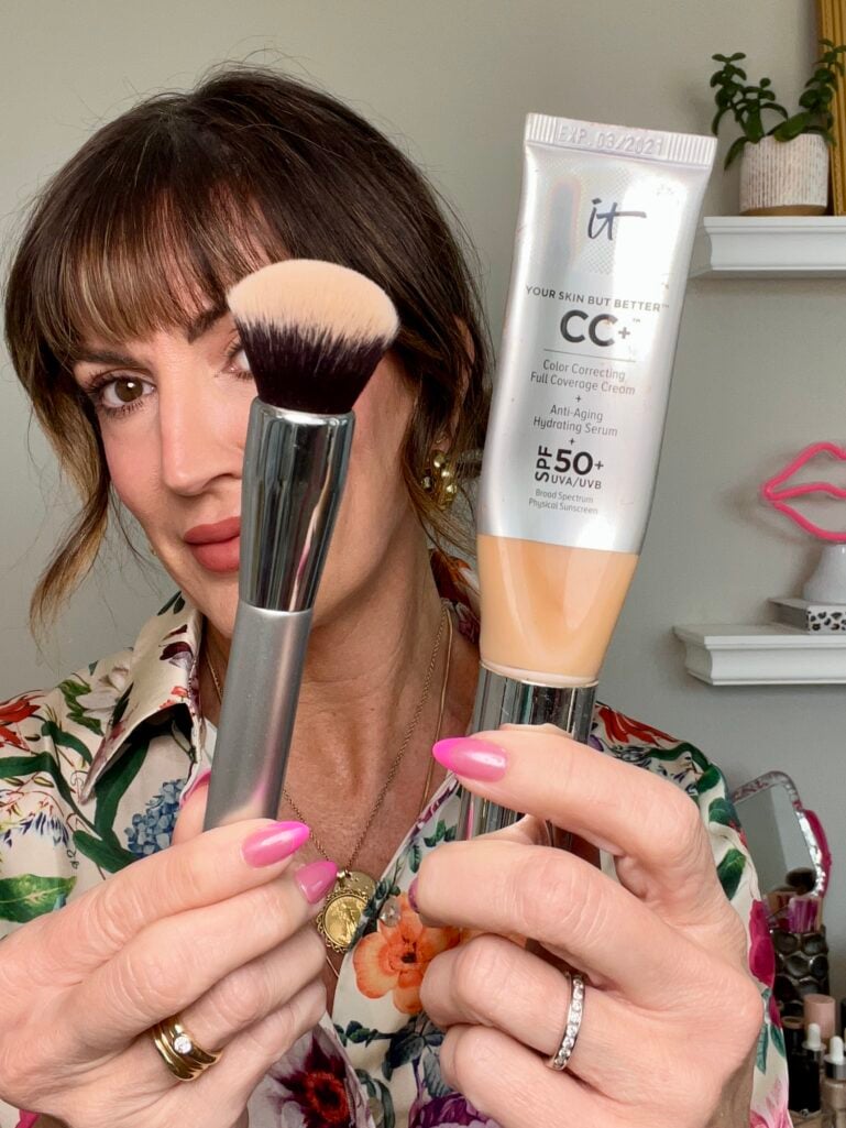 Close-up of woman holding It Cosmetics CC+ Cream SPF 50 tube and It Cosmetics airbrush blurring foundation brush in front of face, floral print blouse, hot pink nails, gold coin pendant necklace and stacking rings, neon pink lip sign on shelf in background