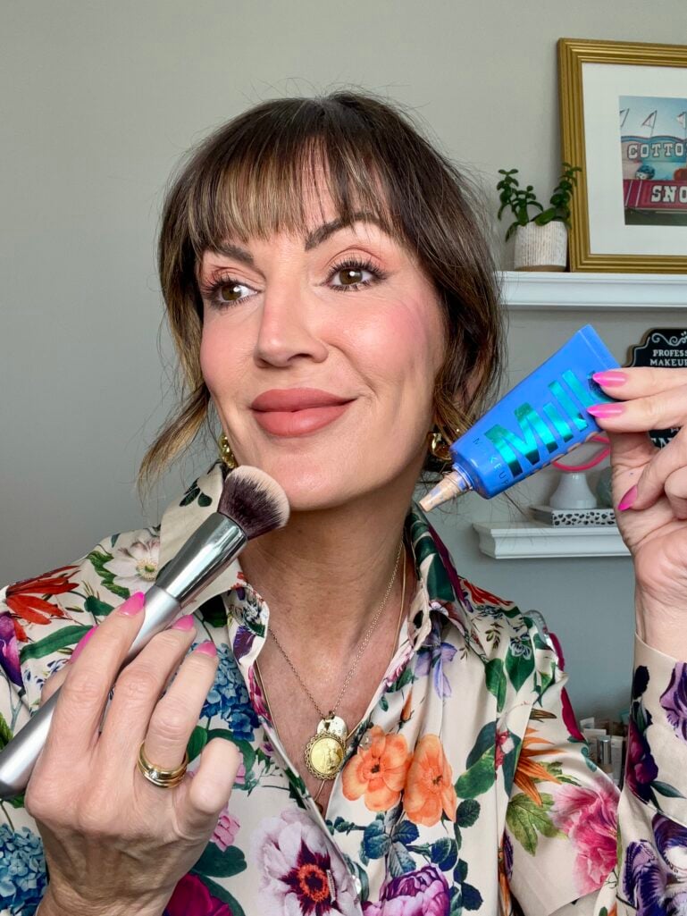 Woman holding blue Milk Makeup Hydro Grip tube and foundation blending brush near face, wearing full Georgia Peach makeup look with warm peach eyes, rosy cheeks, and peachy matte lips, floral button-down blouse, gold chunky hoop earrings and coin necklace, hot pink nails