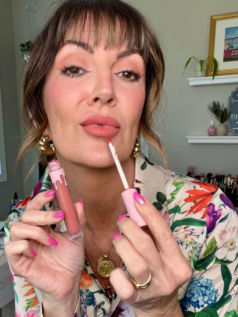 Woman applying Kylie Cosmetics matte liquid lipstick in peach shade with doe-foot applicator, holding tube open near lips, wearing floral print blouse, peach-toned eye and cheek makeup, gold chunky hoop earrings, gold coin necklace and rings, hot pink nails, makeup brushes visible on shelf in background