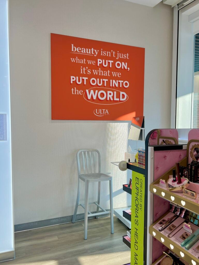nside Ulta Beauty store showing orange wall sign with inspirational quote about beauty, silver metal bar stool beneath, pink tufted display shelving with beauty products, sunlight streaming through store window