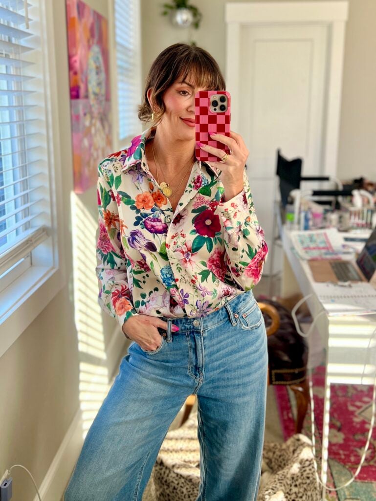Mirror selfie of woman wearing silky ivory floral button-down blouse tucked into light wash wide-leg denim jeans, hair in low updo with curtain bangs, gold chunky hoop earrings and coin pendant necklace, holding phone in pink and red checkered case, bright floral abstract painting on wall, home office desk with laptop visible in background