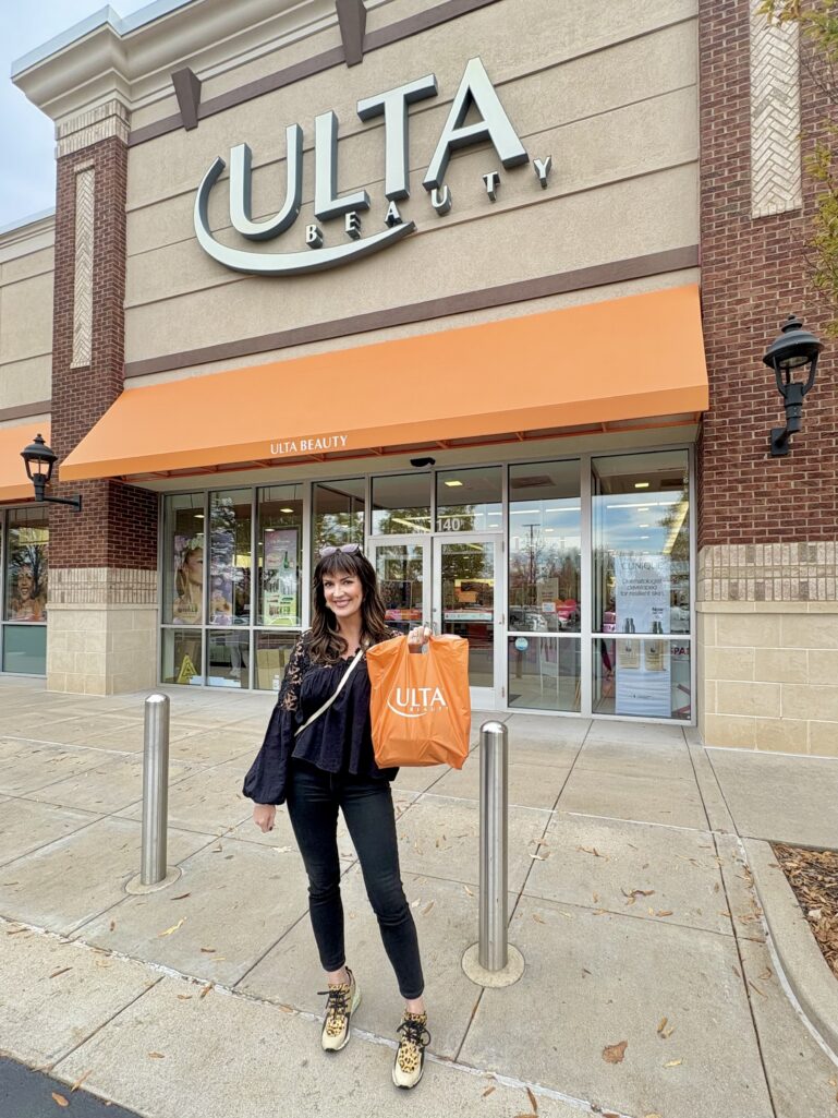 Woman with long dark brown hair and bangs standing in front of Ulta Beauty storefront with orange awning, holding large orange Ulta Beauty shopping bag, wearing black flowy blouse with lace detail, dark skinny jeans, and leopard print sneakers, sunglasses perched on head, smiling at camera