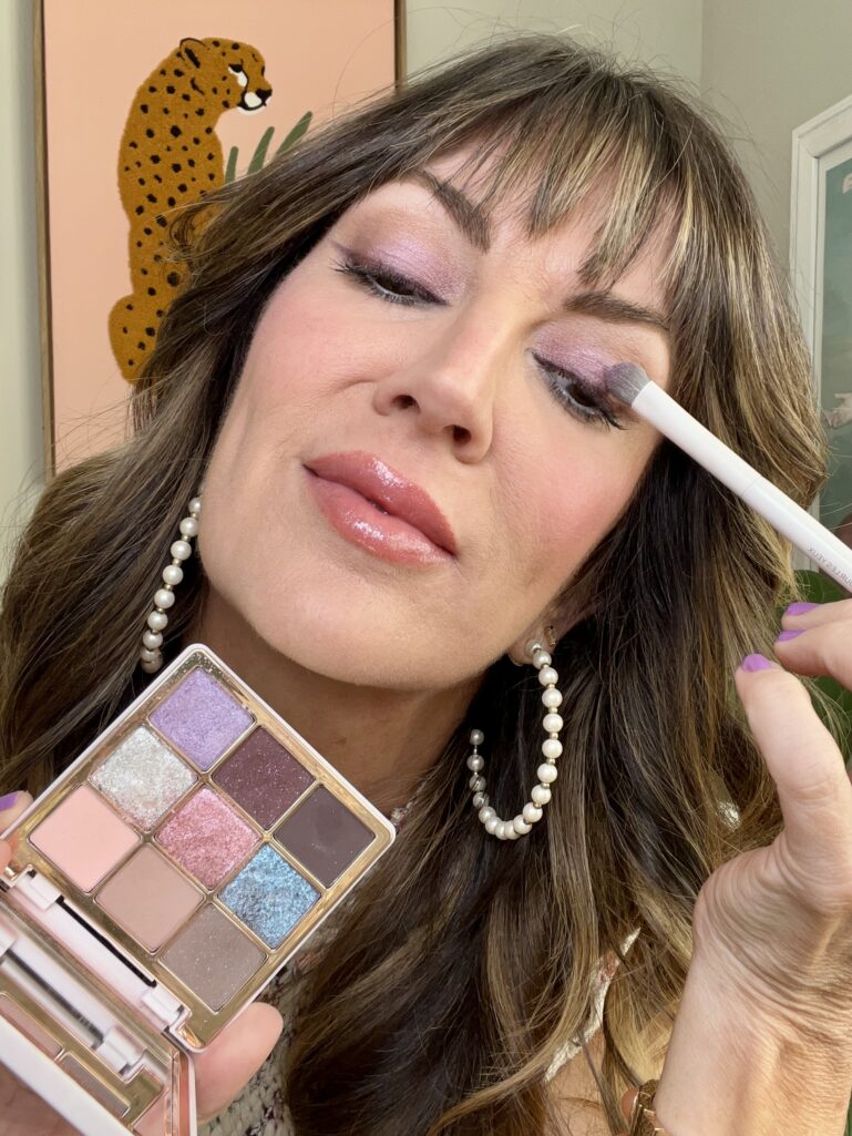 Close-up of woman applying lavender shimmer eyeshadow with white brush while holding Anastasia Beverly Hills mini Sugar palette featuring purple, pink, and blue pastel shades, eyes partially closed showing full lilac eye look, pearl hoop earrings, lavender nails, leopard print wall art in background