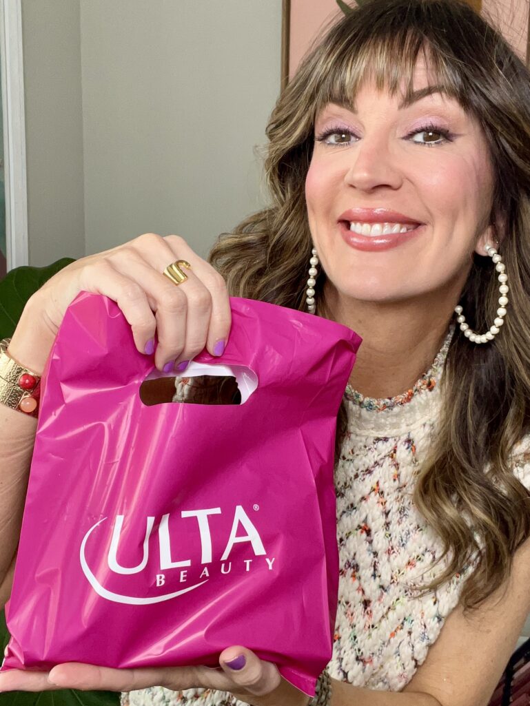 Close-up of woman smiling and holding hot pink Ulta Beauty shopping bag, pearl hoop earrings, cream multicolor tweed sleeveless top, lavender eyeshadow, gold initial ring, colorful gemstone and gold bracelet visible