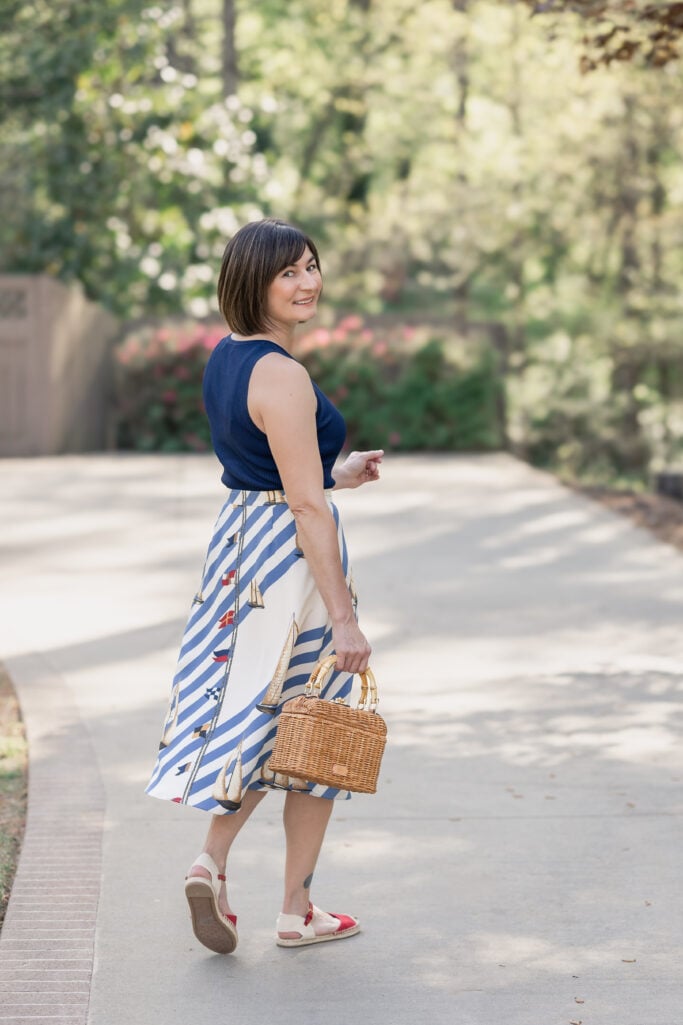 Back view of Kelly in Ralph Lauren nautical sailboat stripe crepe midi skirt with navy ribbed sleeveless top, red canvas espadrilles and wicker lunchbox bag — petite nautical spring outfit
