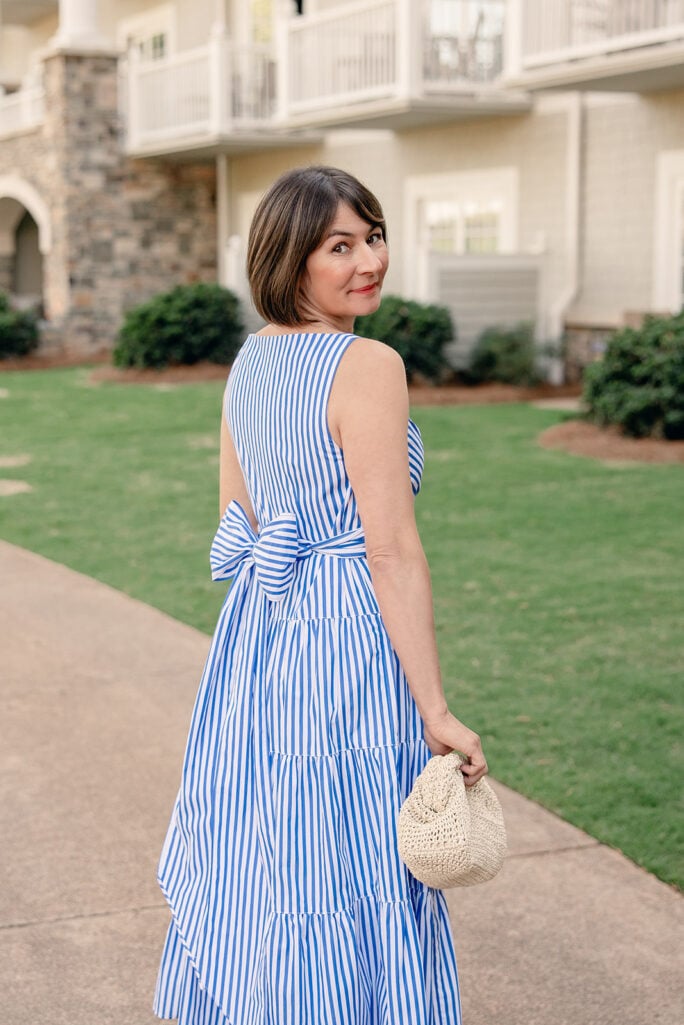 Back view of Kelly in blue and white striped tiered cotton dress showing self-tie bow waist detail and tiered skirt, with straw clutch — petite occasion dress summer
