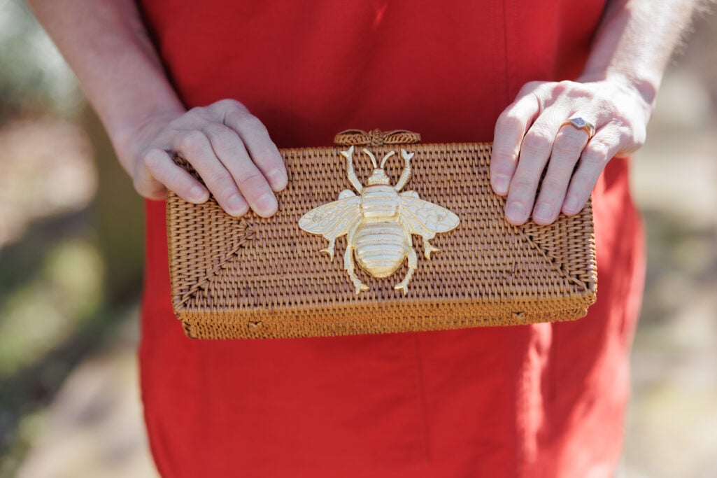 Close-up of handwoven straw clutch with gold bee embellishment held against tomato red dress — spring accessory for petite outfit