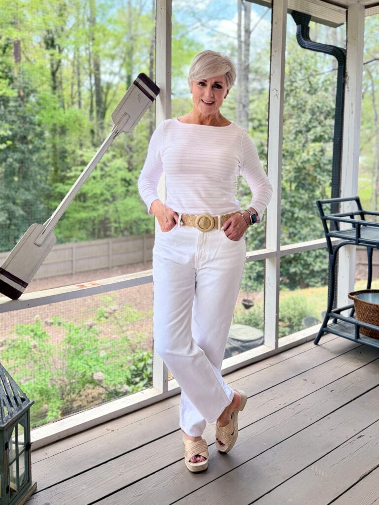 Beth stands on the screened porch in an all-white outfit — a long-sleeve striped tee, white wide-leg denim, a woven straw belt with a round buckle, and raffia platform slide sandals — hands in her pockets, smiling at the camera.