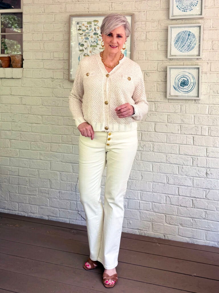 Beth stands on a screened porch against a white brick wall, smiling at the camera, wearing a cream crochet cardigan with gold buttons, ivory bootcut jeans with gold button fly detail, and tan heeled sandals, with a Georgia map print framed behind her.