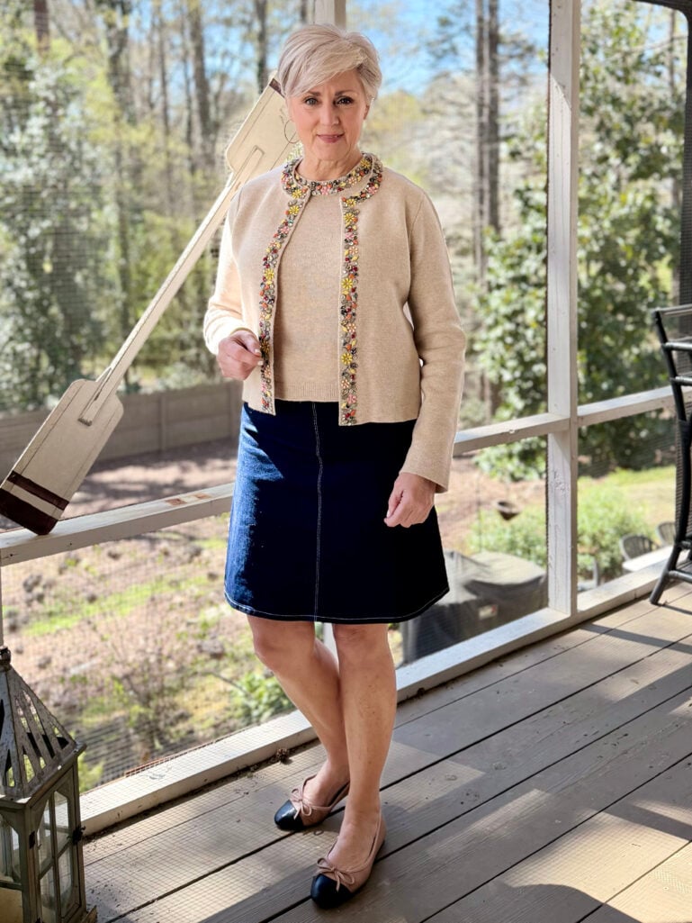 Beth stands on a screened porch wearing a cream embellished knit cardigan over a matching jeweled tee, a dark denim A-line skirt, and two-tone cap toe ballet flats, looking over her shoulder toward the camera.
