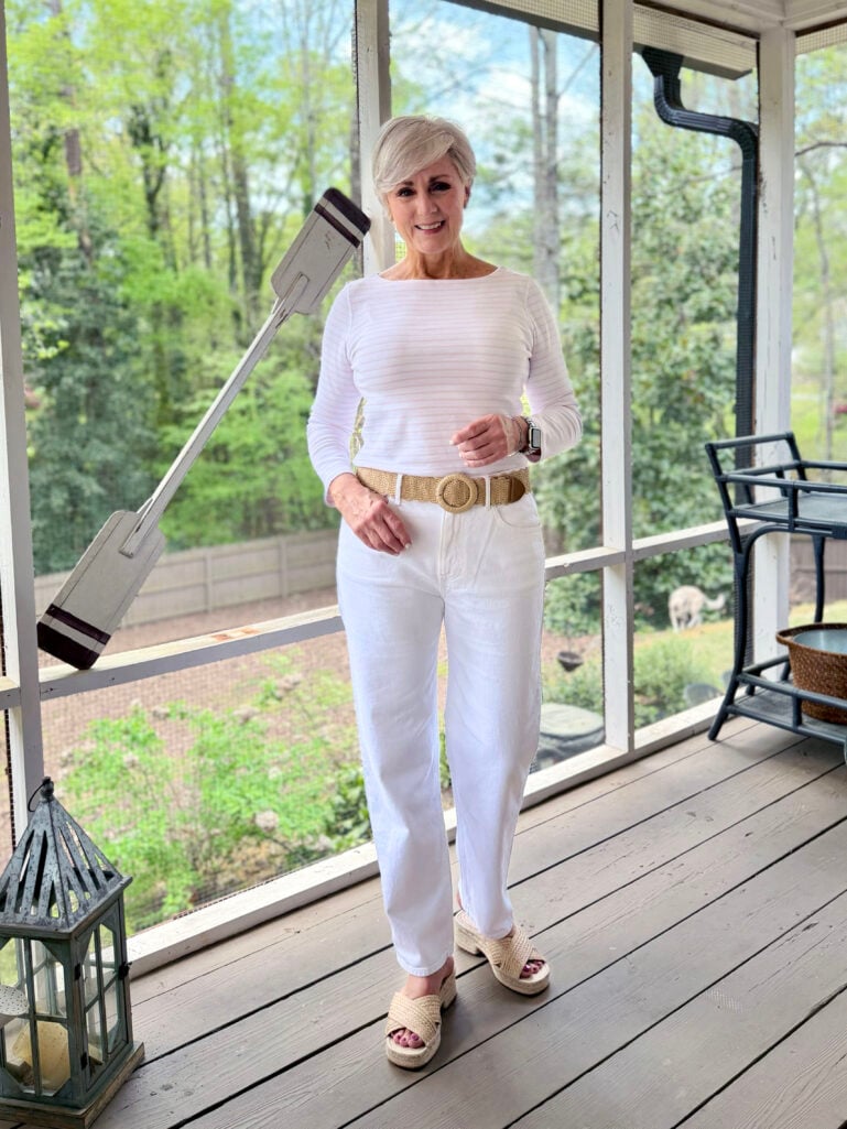 Full-length view of Beth on the screened porch in a white striped long-sleeve tee, white wide-leg jeans, a woven straw belt, and raffia platform slides, smiling warmly with lush green spring trees visible behind her.
