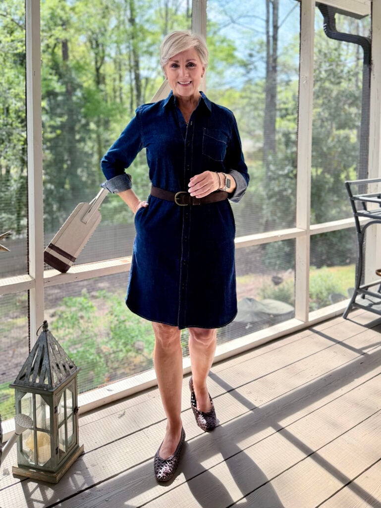 Full-length view of Beth on the screened porch in her belted dark denim shirtdress and woven flats, smiling warmly with lush green spring foliage visible through the screens behind her.