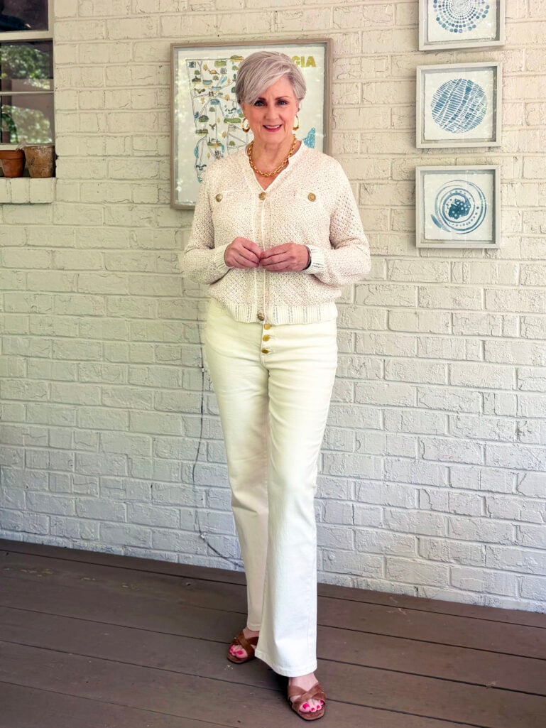 Full-length view of Beth on the porch in her cream knit cardigan, ivory wide-leg jeans, and tan heeled sandals, hands clasped in front, with framed Georgia map art and blue abstract prints visible on the white brick wall behind her.