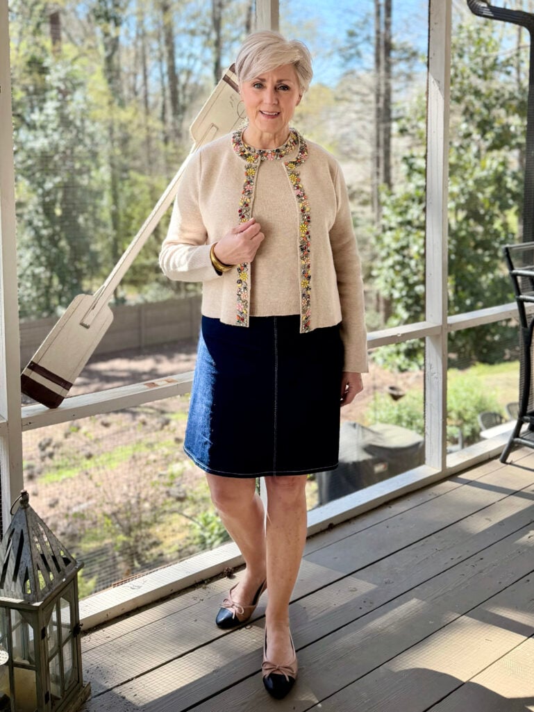 Full-length view of Beth on the screened porch in a cream embellished cardigan and tee set, dark denim skirt, and nude and black cap toe flats, smiling at the camera with one hand at her chest.