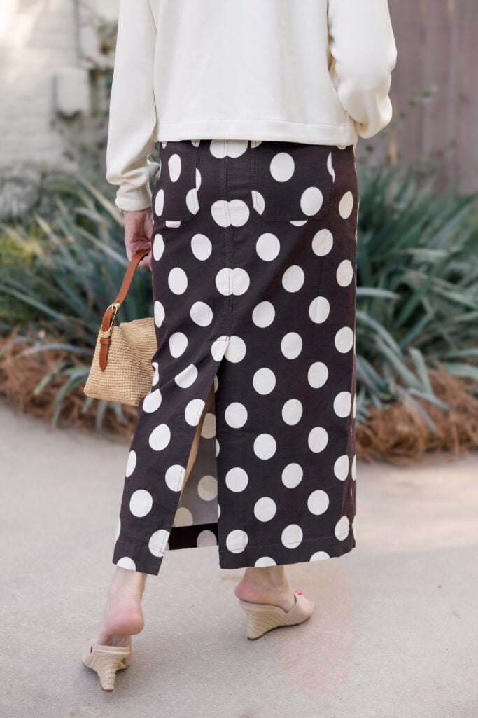 style has no expiration date — woman over 50 in a black and white polka dot maxi skirt, charcoal tee, ivory cropped jacket, gold chain necklace and hoops, straw bucket bag, and nude espadrille wedge sandals