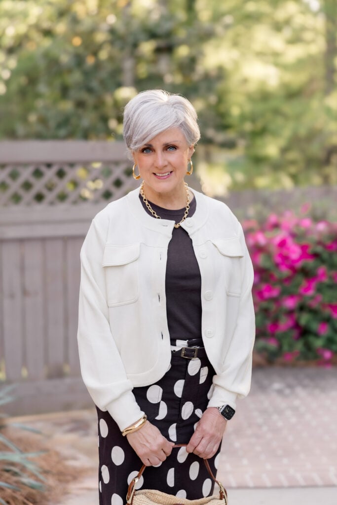  personal style over 60 — woman in a brown and white polka dot maxi skirt and ivory jacket walking confidently outdoors, garden and wooden fence in the background