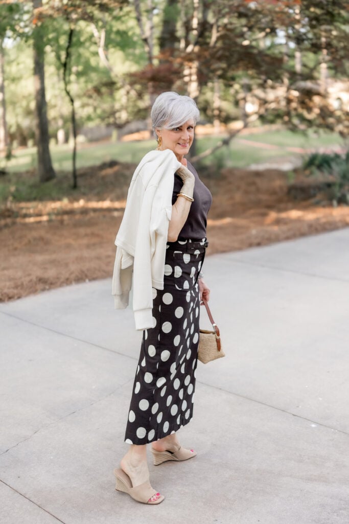 women over 50 with style — woman with silver hair smiling in an ivory cropped jacket and brown and white polka dot maxi skirt, holding a straw bucket bag, golden hour outdoor light