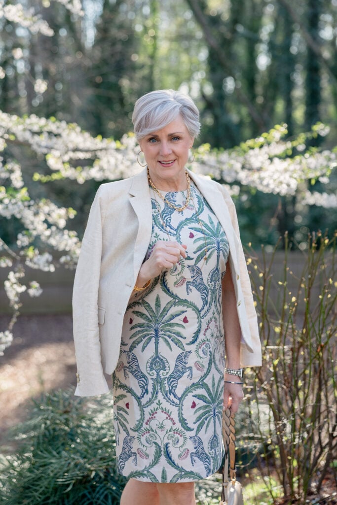 Woman over 50 with silver hair walking a stone garden path in spring, wearing a tropical print shift dress with palm trees and zebras in green, blue, and pink tones, layered under a neutral linen blazer. She carries a structured taupe Tory Burch shoulder bag with gold hardware and wears nude sandals, a pearl and gold layered necklace, and gold hoop earrings — a polished Easter accessories look for women over 50.
