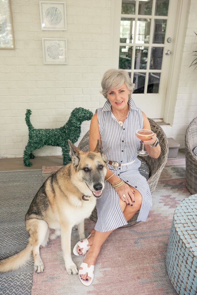 Beth sits in a wicker chair on a covered patio in a summer striped dress, her arm around Oscar, her German shepherd, who leans contentedly against her. A sculptural topiary dog is visible in the background.