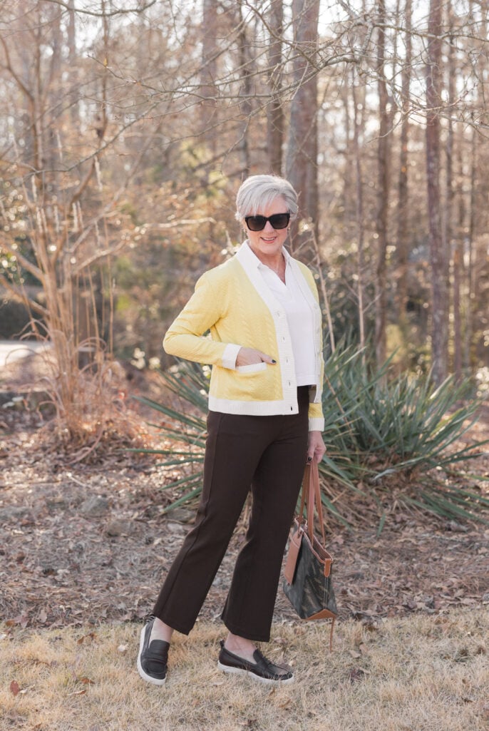 Woman over 50 with silver hair wearing a butter yellow cable-knit cardigan with ivory trim over a white collared shirt, styled with chocolate brown straight-leg trousers, black leather slip-on sneakers, black sunglasses, and a brown leather tote bag — a warm and wearable butter yellow spring look for women over 50.