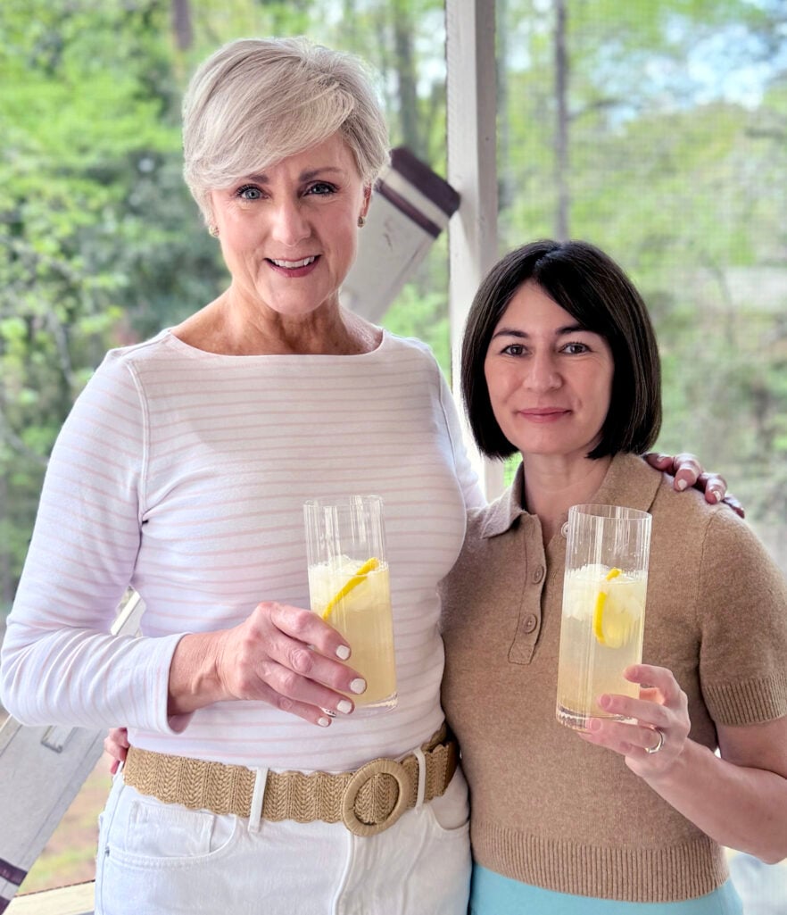 Beth and Kelly stand together on the screened porch, each holding a tall Elderflower Collins cocktail garnished with a lemon twist — Beth in white with a straw belt, Kelly in a camel polo sweater — both smiling at the camera.