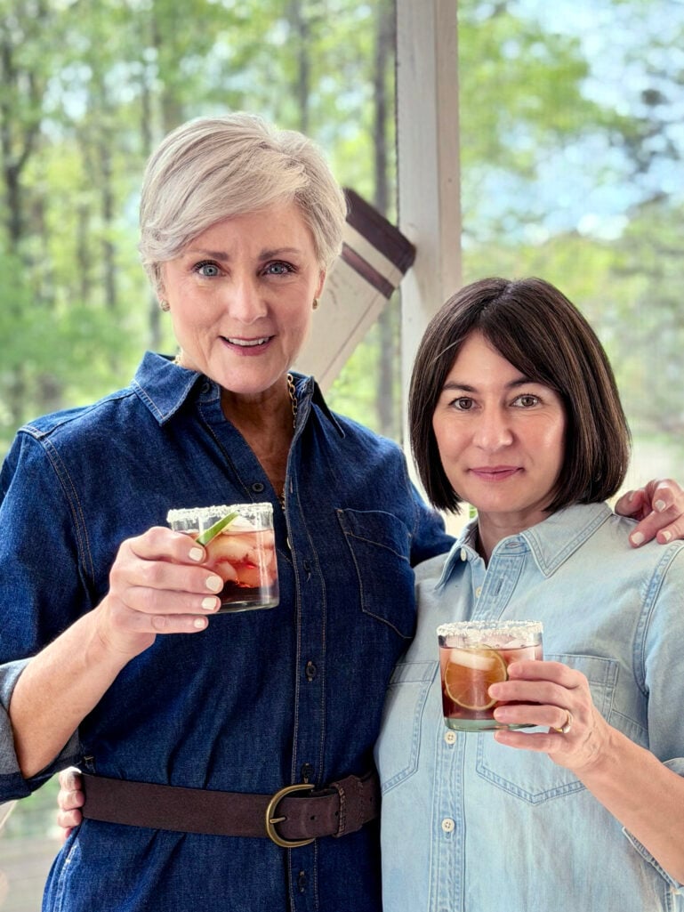 Beth and Kelly stand together on the screened porch, each holding a deep red Hibiscus Margarita in a salted-rim rocks glass garnished with a lime wheel — Beth in her dark denim shirtdress, Kelly in her light chambray cropped top.