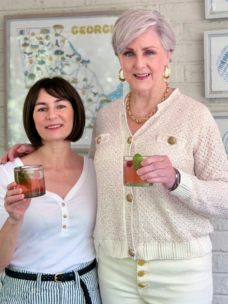 Kelly and Beth stand close together on the porch, each holding a rocks glass filled with rosy pink berry sangria garnished with a raspberry and mint sprig, smiling at the camera.