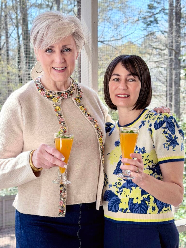 Beth and Kelly stand together on the screened porch holding Peach Bellinis in champagne flutes, both smiling at the camera — Beth in her cream embellished cardigan, Kelly in her blue and yellow floral sweater tee.