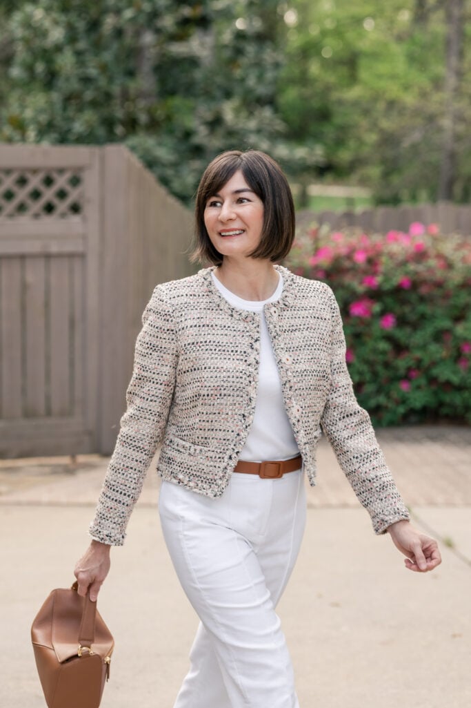 Kelly in three-quarter length shot, walking toward the camera, wearing cream tweed lady jacket over white tee with cognac belt and leather bag — petite spring polished outfit