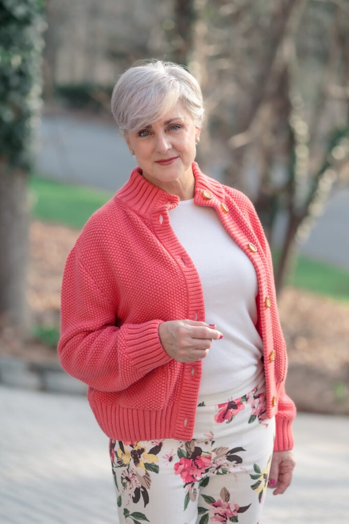 Woman over 50 with silver hair wearing a coral chunky-knit button-front sweater jacket over a white fitted cotton tee, paired with cropped floral slim pants in cream with pink and green blooms — a warm, spring-ready coral look for women over 50 that coordinates color through print.