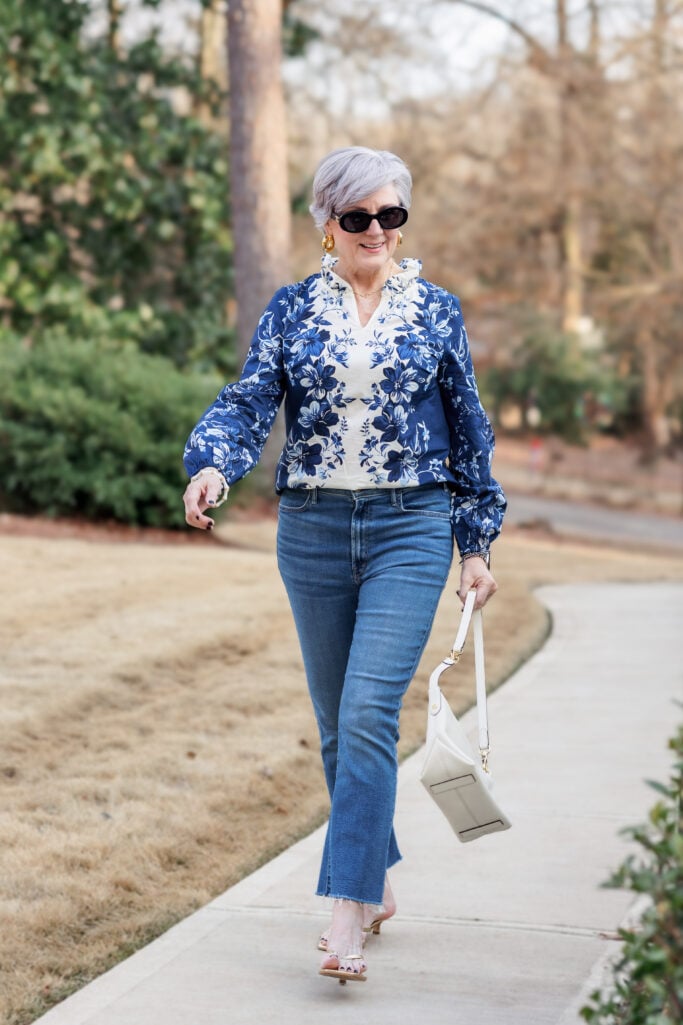 Woman over 50 with silver hair walking confidently in a navy and ivory floral print top with ruffle neckline detail, paired with medium-wash flare jeans, a cream structured mini shoulder bag, gold hoop earrings, oval black sunglasses, and gold kitten heel thong sandals — a chic casual cobalt blue look for spring over 50.