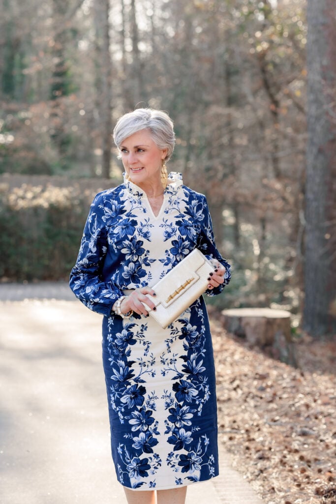 Woman over 50 with silver hair wearing a navy and ivory floral print shift dress with a mandarin collar and blouson sleeves, holding a gold bamboo-detail clutch and wearing gold drop earrings and a pearl necklace — a polished example of how to wear cobalt and navy for spring over 50.
