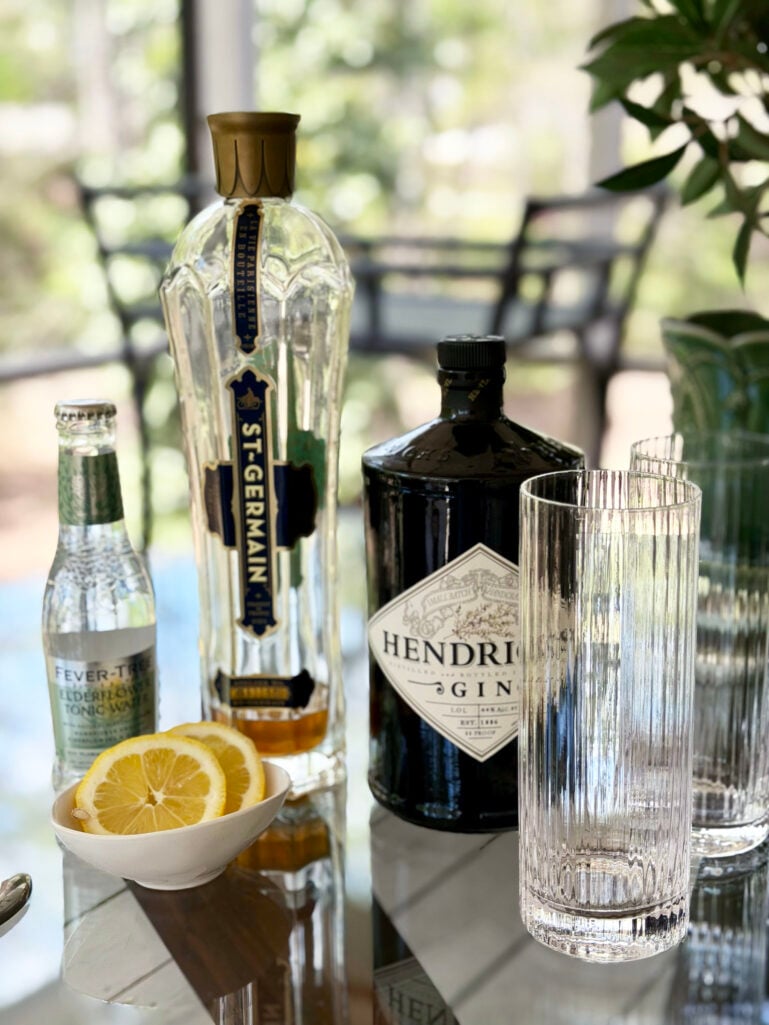 A flat lay of Elderflower Collins ingredients on a glass table: a bottle of St-Germain elderflower liqueur, a bottle of Hendrick's Gin, a small bottle of Fever-Tree Elderflower Tonic Water, two empty ribbed highball glasses, and a small bowl of sliced lemons.