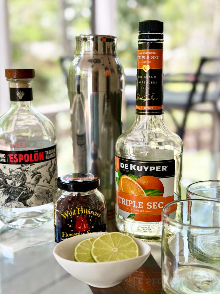 A flat lay of Hibiscus Margarita ingredients on a glass table: a bottle of Espolón Blanco tequila, a jar of Wild Hibiscus Flowers in Syrup, a bottle of De Kuyper Triple Sec, a silver cocktail shaker, two rocks glasses, and a small bowl of sliced limes.