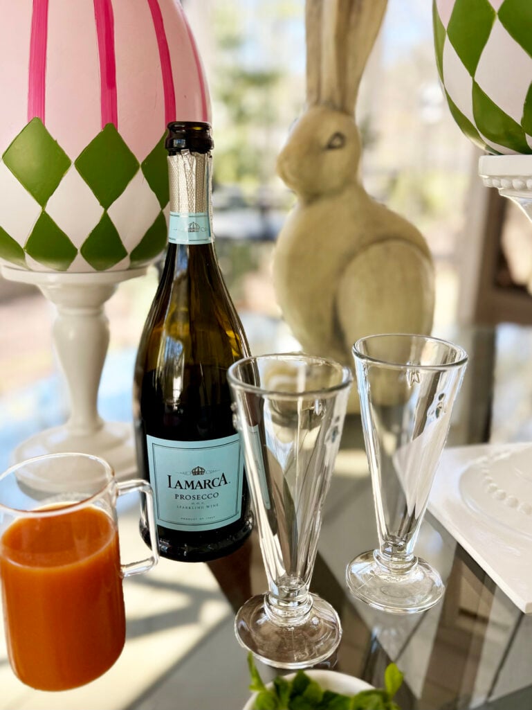 A bottle of La Marca Prosecco, a small glass pitcher of peach nectar, two empty champagne flutes, and a small bowl of fresh mint sit on a glass table surrounded by colorful Easter egg and bunny decorations.