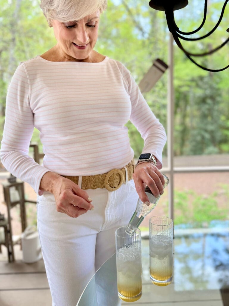 Beth pours St-Germain elderflower liqueur into two ice-filled ribbed highball glasses already holding lemon juice, wearing her white striped tee and woven straw belt, smiling down at the cocktails.