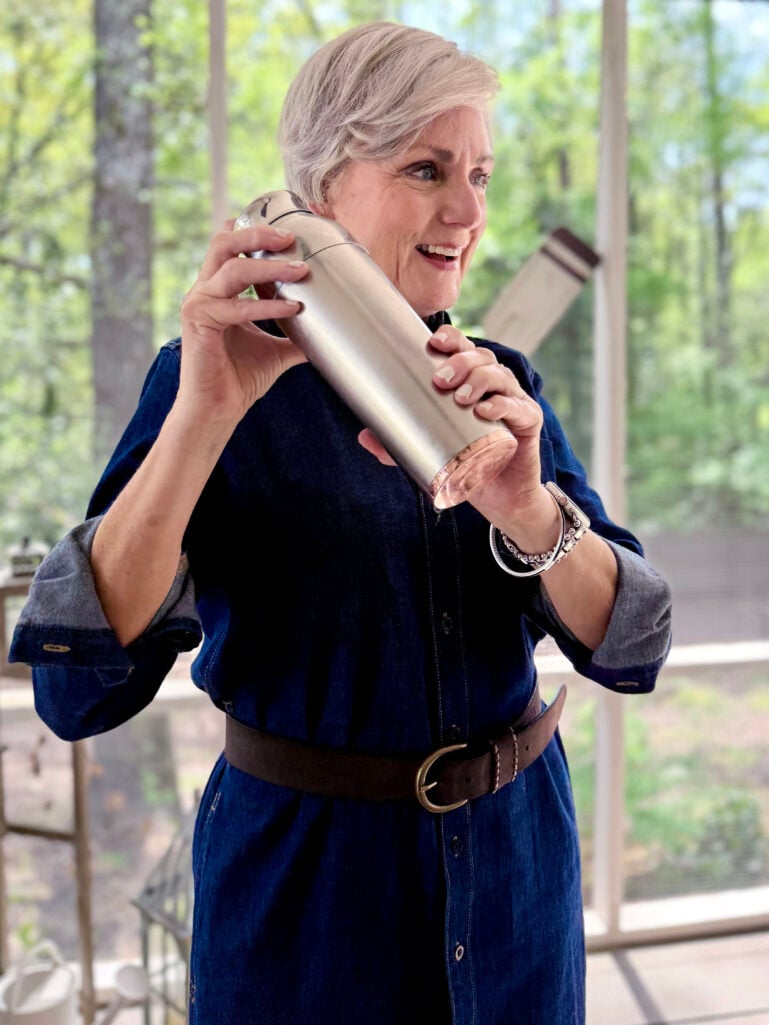 Beth laughs while vigorously shaking a silver cocktail shaker on the screened porch, wearing her belted dark indigo denim shirtdress, lush green spring trees visible behind her.