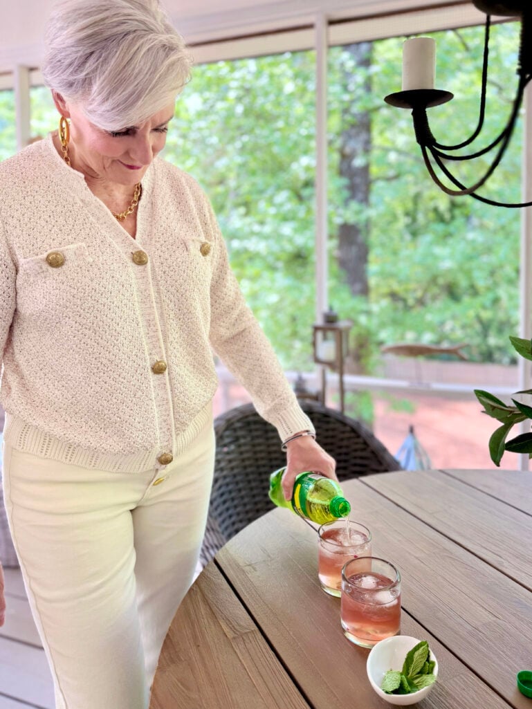 Beth leans over an outdoor table, pouring ginger ale from the bottle into two rocks glasses filled with pale pink berry sangria, a small white bowl of mint nearby, with the screened porch and garden visible behind her.