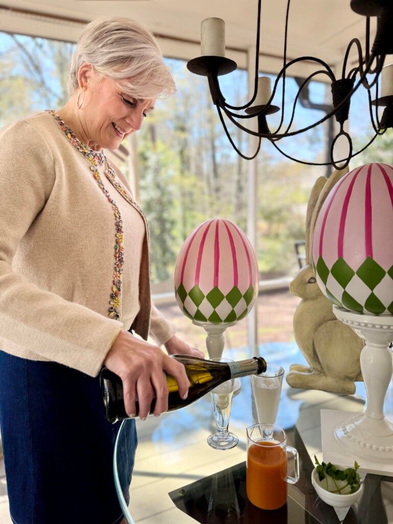 Beth pours Prosecco from a bottle into two champagne flutes already filled with peach nectar, smiling as she prepares the Bellinis beside festive pink and green Easter egg decorations and a ceramic bunny.